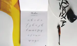 Na stronie z alfabetem widać, jak się pisze duże T. Obok leżą stalówki i kałamarz.