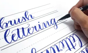 Ręczne pisanie liter, czyli hand lettering, w kolorze niebieskim na papierze w linie. Dłoń trzyma pisak.