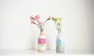 Dwa urocze wazony DIY z butelek, ozdobione sznurkiem i filcowymi kwiatami, z różowymi i żółtymi goździkami.