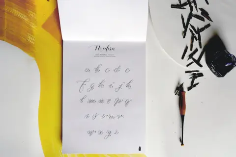 Na stronie z alfabetem widać, jak się pisze duże T. Obok leżą stalówki i kałamarz.