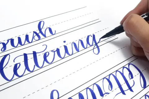 Ręczne pisanie liter, czyli hand lettering, w kolorze niebieskim na papierze w linie. Dłoń trzyma pisak.