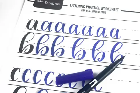 Ćwiczenie kaligrafii litery "a" i "b" na arkuszu Tombow. Niebieski pisak i jego nakrętka leżą obok.