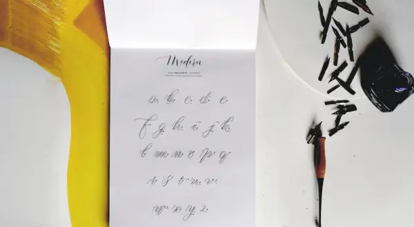 Na stronie z alfabetem widać, jak się pisze duże T. Obok leżą stalówki i kałamarz.