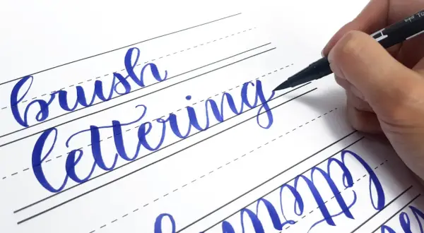 Ręczne pisanie liter, czyli hand lettering, w kolorze niebieskim na papierze w linie. Dłoń trzyma pisak.