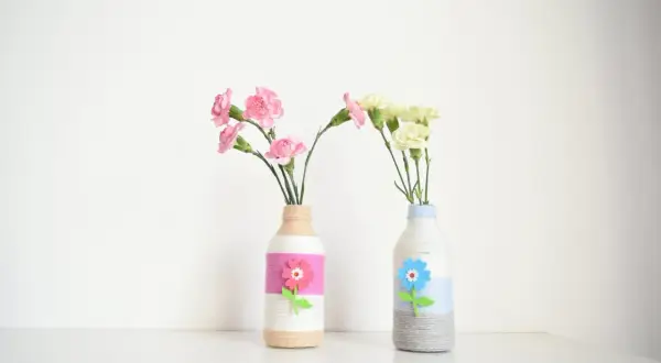 Dwa urocze wazony DIY z butelek, ozdobione sznurkiem i filcowymi kwiatami, z różowymi i żółtymi goździkami.