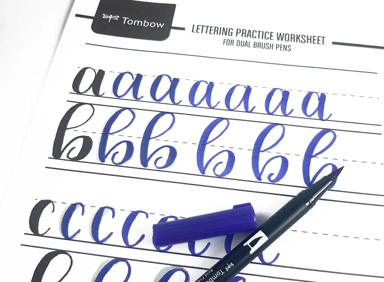 Ćwiczenie kaligrafii litery "a" i "b" na arkuszu Tombow. Niebieski pisak i jego nakrętka leżą obok.