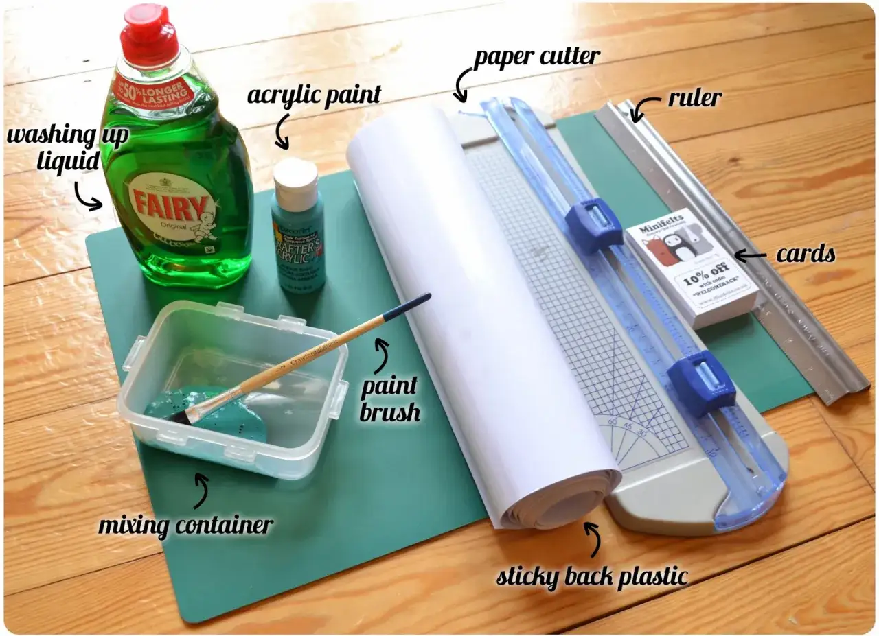 Potrzebne materiały, by zrobić zdrapkę: płyn do naczyń, farba akrylowa, pędzel, pojemnik, papierowa mata, nożyczki, linijka, karty i folia samoprzylepna.