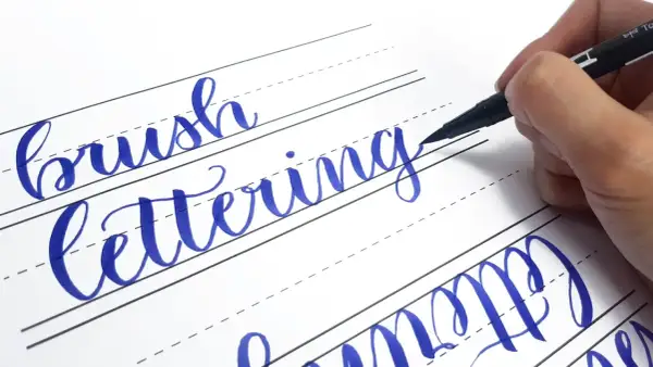 Ręczne pisanie liter, czyli hand lettering, w kolorze niebieskim na papierze w linie. Dłoń trzyma pisak.